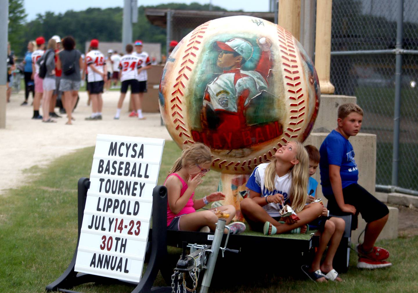 Children munch on snacks near a large baseball statue during MCYSA 2023 Summer International Championships Opening Ceremonies Friday, July 14, 2023, at the Mickey Sund Complex in Lippold Park in Crystal Lake.