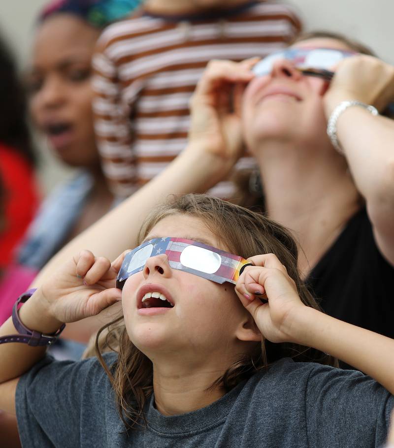Katie Krupa of DeKalb (front), 9, and her mother Heather Krupa (back right) try to find the sun through the clouds using solar eclipse glasses on Monday, Aug. 21, 2017 at the DeKalb Public Library in DeKalb. Heather purchased the glasses for $1 each at Walmart a few weeks ago.