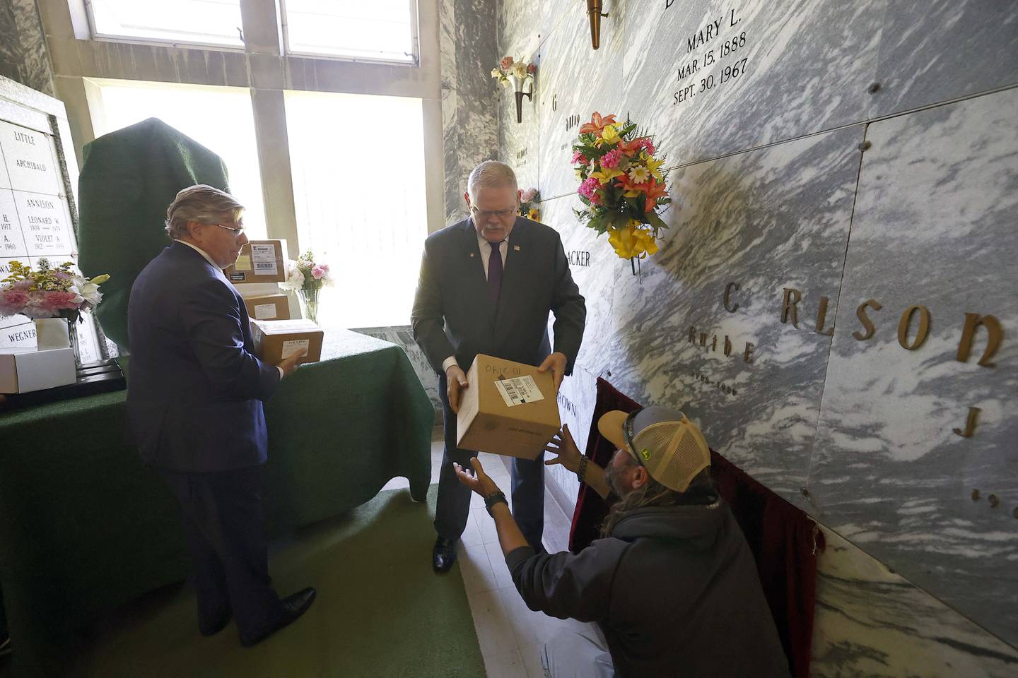 Funeral Director of Yurs Funeral Homes Timothy Bergman, left, and Kane County Coroner Rob Russell, center, take turns handing boxes of cremains to St. Charles Township North Cemetery Superintendent Brooks Ronzheimer who then places them in a crypt during a ceremony for the entombment of unclaimed bodies Thursday May 30, 2024 in St. Charles.