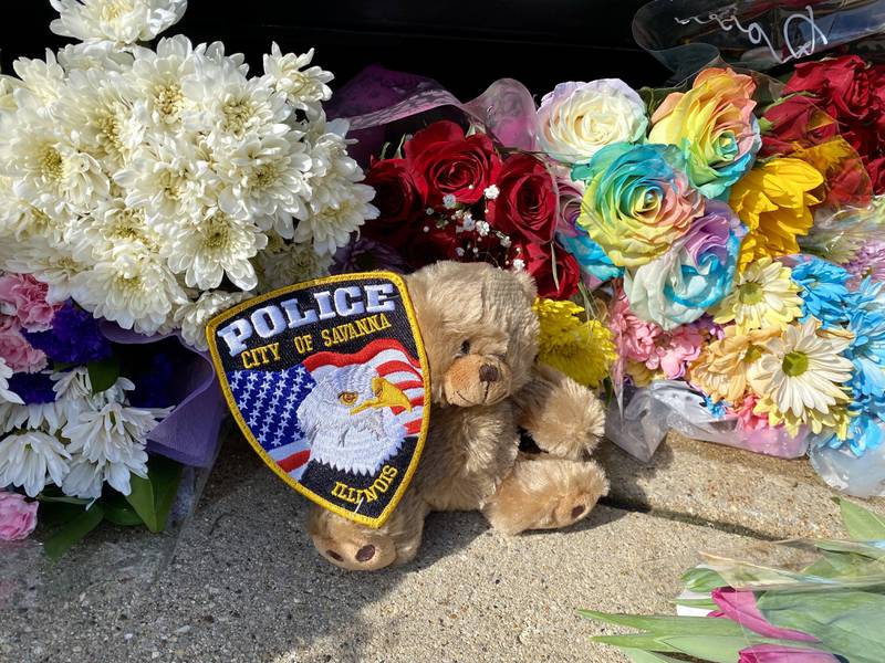 A City of Savanna Police Department badge sits among dozens of flower arrangements placed near a DeKalb County Sheriff's Office vehicle on the DeKalb County Courthouse lawn Saturday, March 30, 2024. Mourners have visited the site to leave trinkets, flowers and other tokens in memory of sheriff's deputy Christina Musil, who was killed while on duty after her squad car was rear-ended by a truck Thursday, March 28, 2024.