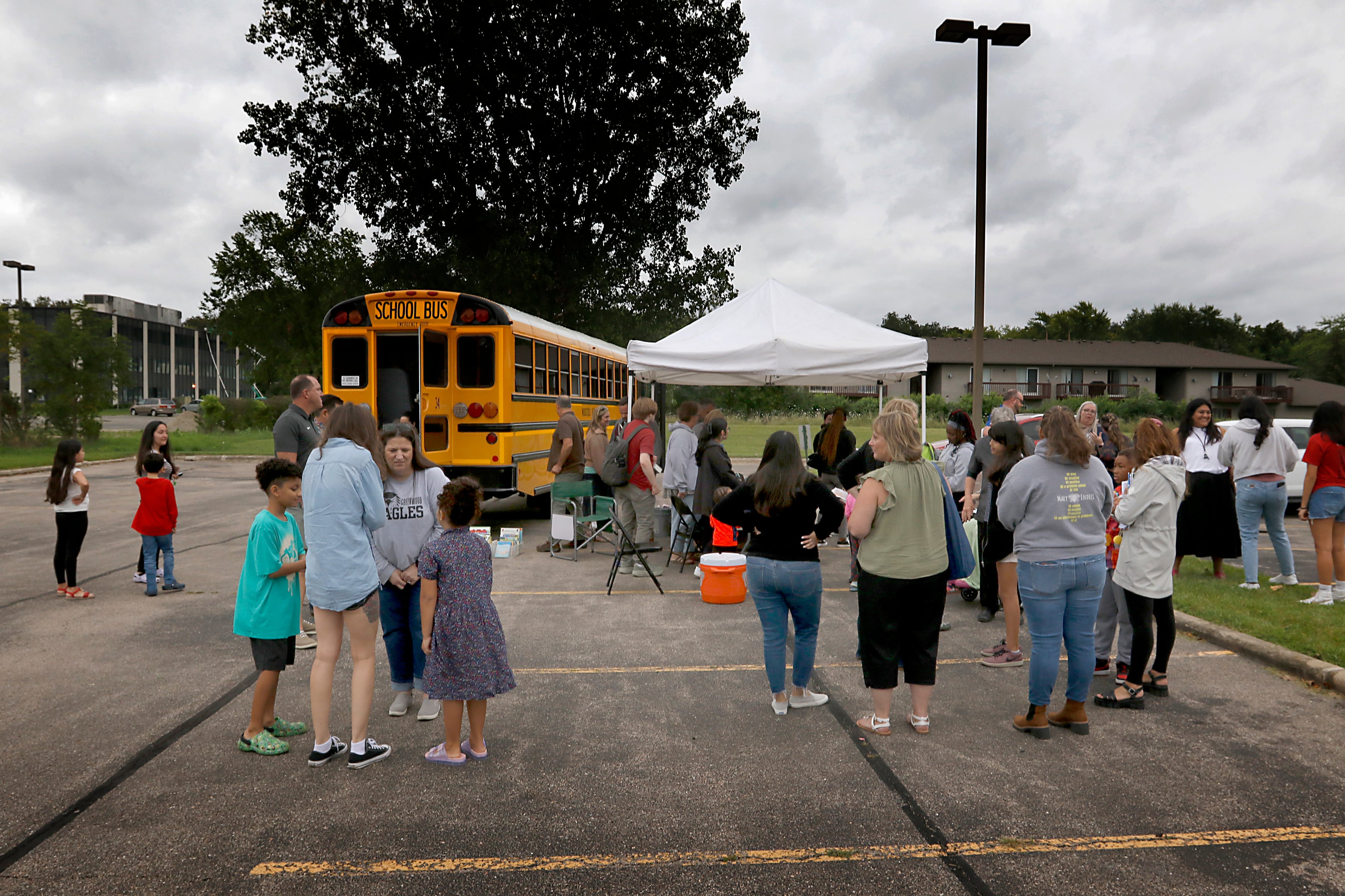 People gather around a school bus during a Woodstock School District 200 Back to School Coming to You event at Sheila Street Apartments on Tuesday, Aug. 6, 2024. The location was one of twelve stops on the tour that gave out backpacks and school supplies, provided registration help and computer repair.