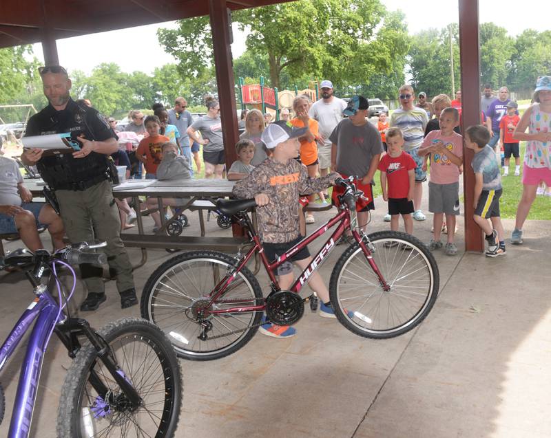 Briggs Moser, 7, of Prophetstown, wheels the new bike he won out of the shelter during the 18th annual Dick Brown Fishing Derby at Prophetstown State Park on Saturday, June 15, 2024. Also pictured is Prophetstown Police Officer Will Lakehart.