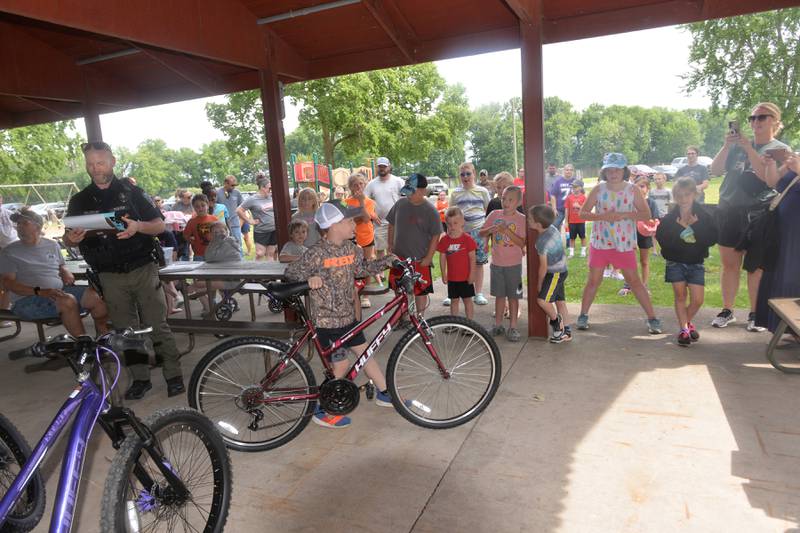 Briggs Moser, 7, of Prophetstown, wheels the new bike he won out of the shelter during the 18th annual Dick Brown Fishing Derby at Prophetstown State Park on Saturday, June 15, 2024. Also pictured is Prophetstown Police Officer Will Lakehart.