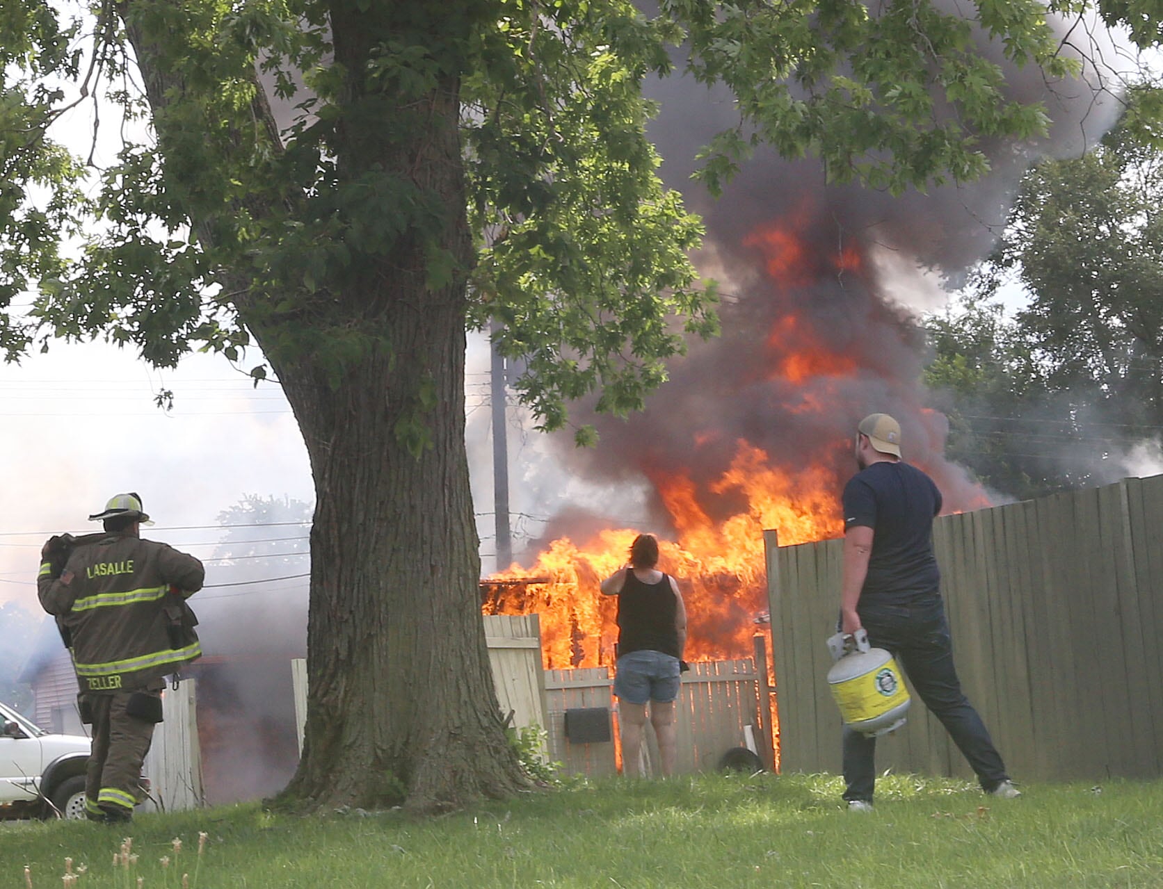 La Salle firefighter Brian Zeller (left) throws on his bunker gear to fiight a fire as neighbors remove propane tanks whlie flames engulf a garage in the 800 block of Lafayette Street on Monday, July 22, 2024. The fire began just before 1p.m. La Salle Fire and EMS along with Peru Fire department responded to the call while La Salle Police directed traffic.