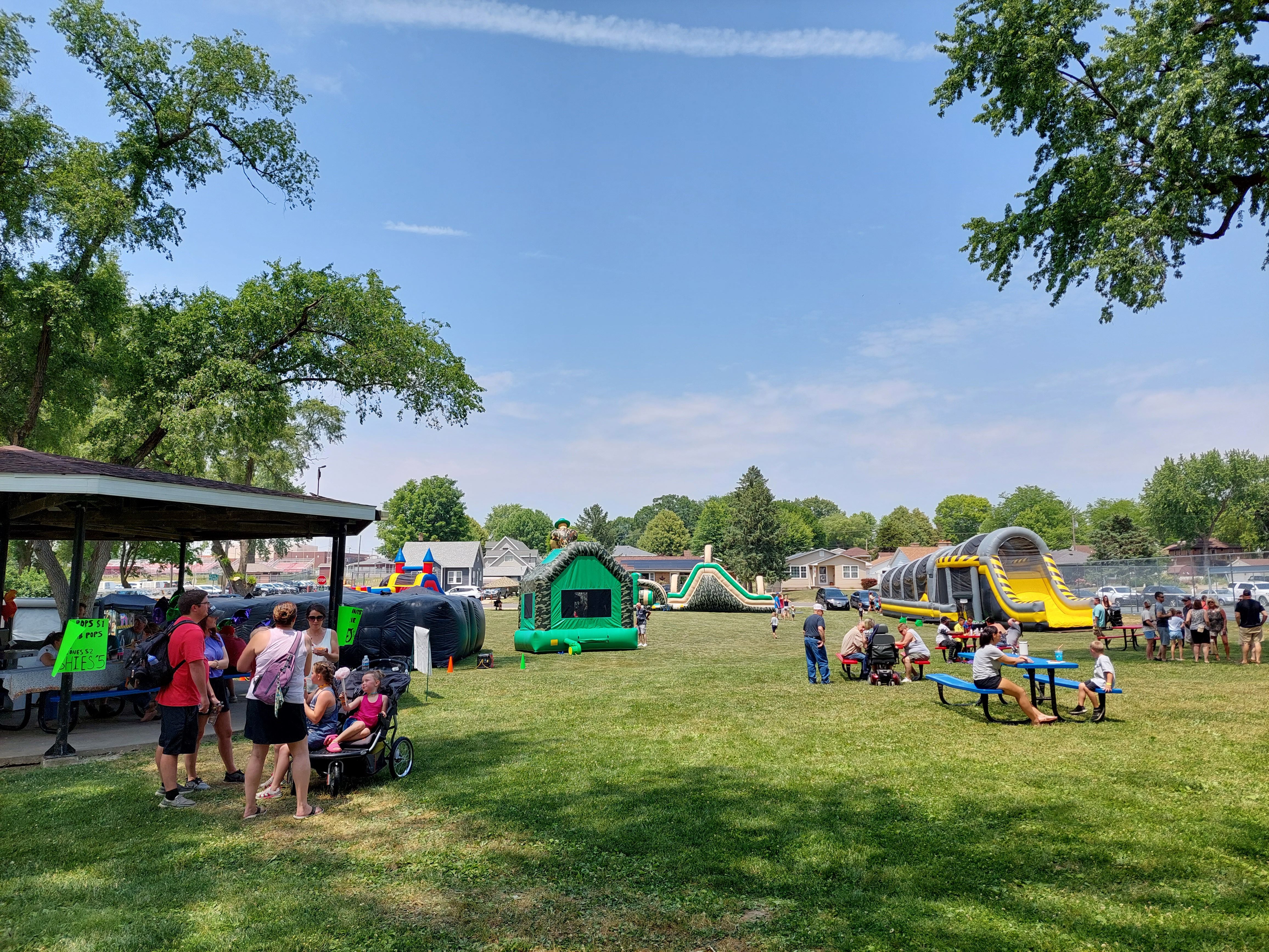 Summerfest attendees sit at picnic tables as children run through a number of inflatable bounce houses and obstacle courses Saturday, June 10, 2023, during Summerfest at Kirby Park in Spring Valley.