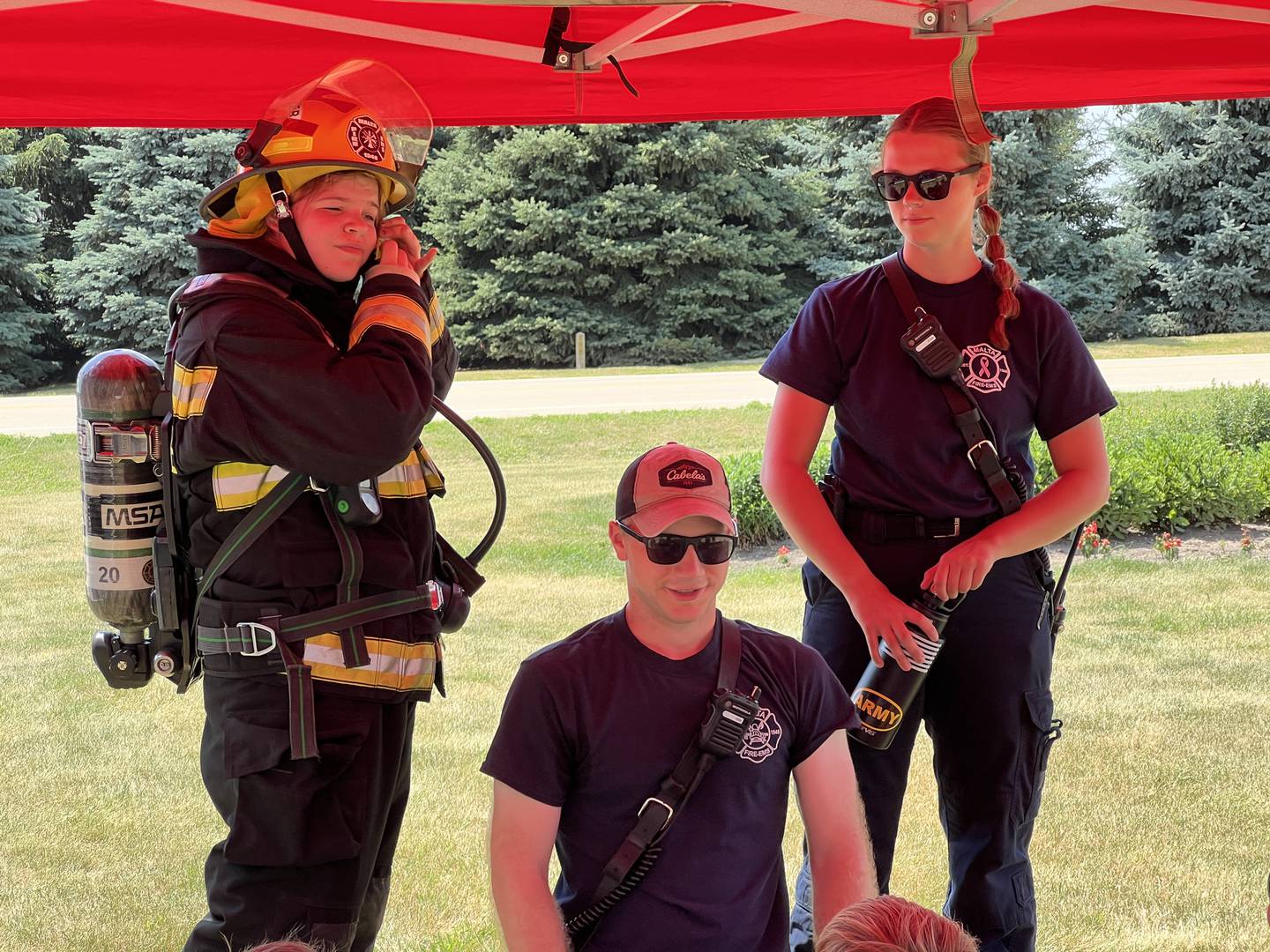 Addison McGlaughlin, wearing protective gear alongside Sam Wurtz and Sydney Thorne talk to farm safety campers on June 19, 2024 about fire safety – and how heavy a firefighter's equipment is – for the Malta Fire Protection District.