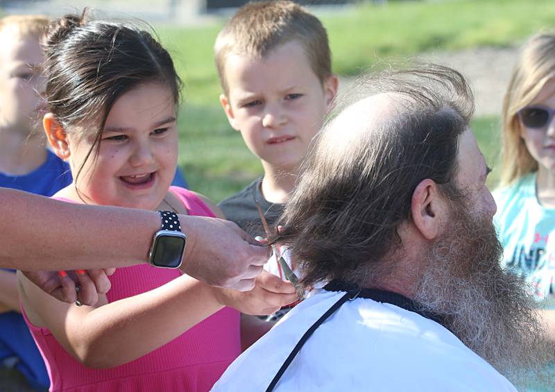 Estella, a student in Mrs. Richards class reacts as she trims a piece of hair off of custodian Bob Espel during his head and beard shaving on Wednesday, May 22, 2024 at Douglas Elementary School in Princeton. Students raised more than $3.332 in the past few weeks. All proceeds will go to the new landscaping around the Douglas building, with the first round of plantings to take place with students present this Thursday, May 23. Using funds from the Preschool for All grant the school has purchased new age-appropriate playground equipment to promote physical activity, creativity and social interaction.