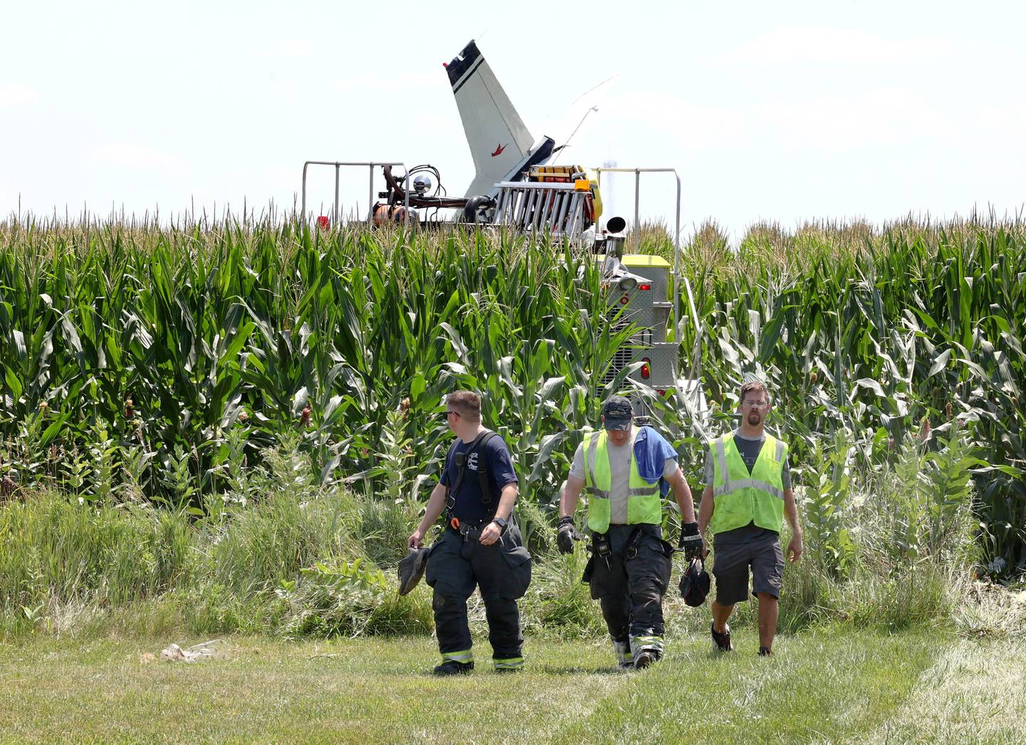 The tail of a plane sticks out above the corn as first responders and airport personnel walk out of the field after responding to a crash Thursday, July 27, 2023, behind a business at 260 West Lincoln Highway in Cortland.