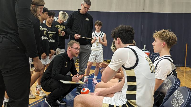 Sycamore coach Ethan Franklin (crouched left) talks to his Spartans during a timeout Wednesday, February 21, 2024 in a Class 3A Belvidere North Regional semifinal against Woodstock North.
