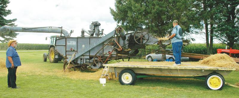 Gerald Van Dusen, of Polo, Illinois, pitches a forkful of wheat straw into Harold Goeking’s threshing machine while Goeking, also of Polo, watches at the 2019 Farm Heritage Festival at East Jordan United Methodist Church.