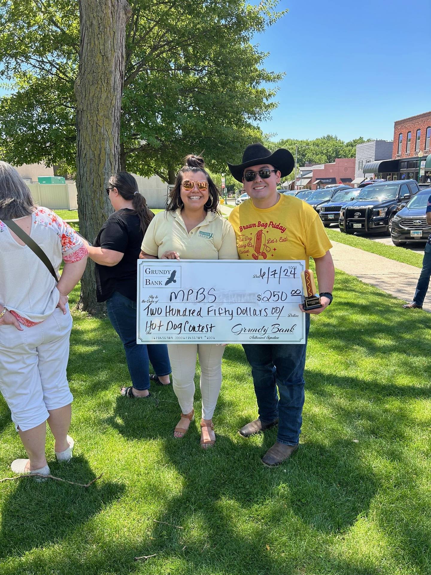 Christina Mendez (left) from Grundy Bank presents Officer Alex Hernandez and his cowboy hat with $250 for winning the 2024 Grundy Bank Hot Dog Eating Contest.