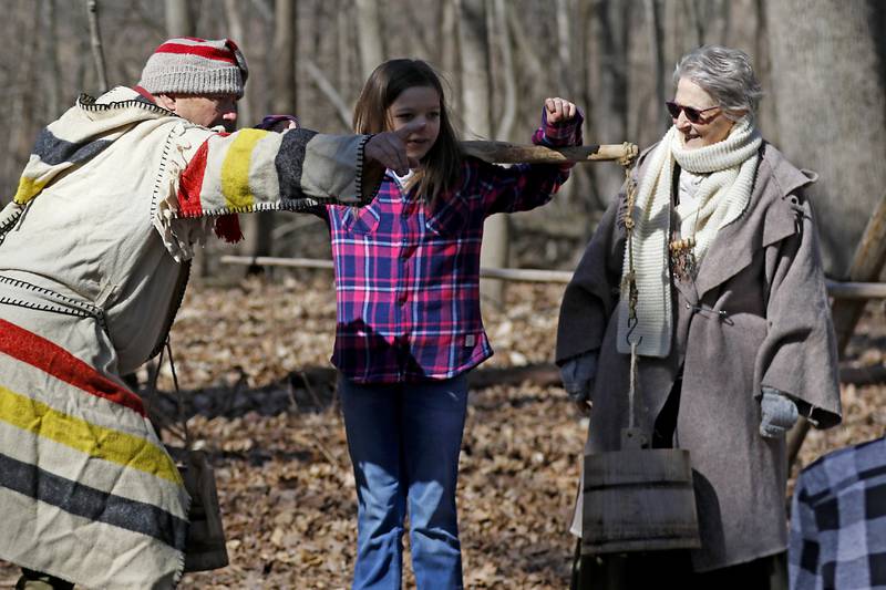 Volunteers Rich Tobiasz and Valerie Koehn explains how much walking and carrying a child would have to do in the early days to Chloe Meyer, 12, of Union and she tries on the yoke and buckets used to collect maple sap during the McHenry County Conservation District’s annual Festival of the Sugar Maples, at Coral Woods Conservation Area, 7400 Somerset Drive in Marengo. People were able to walk through the woods and learn about history of maple sugaring and the evolution of the sap collection process during the one hour long half-mile hike through a woodland trail, on Monday, March 11, 2024. The festival continues on March, 16, and 17.